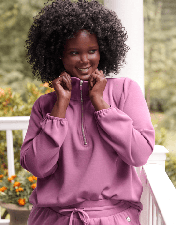 A smiling woman with curly hair wearing a purple tracksuit stands on a white porch against a blurred green background.