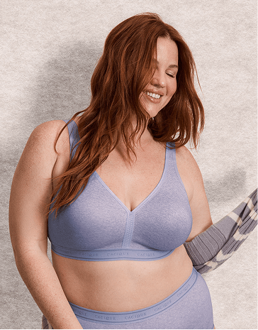 A smiling woman with red hair wearing a light purple lingerie set stands against a neutral, textured grey background.