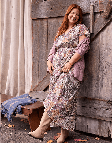 A smiling woman with red hair wearing a patterned dress and purple cardigan leans against a rustic wooden barn door next to a bench with a blue blanket.