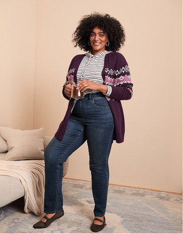 A smiling woman with curly hair wearing a purple cardigan and blue jeans stands in a room with a neutral-colored wall and a beige sofa.