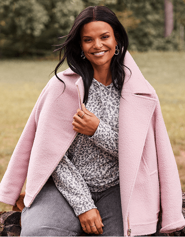 A smiling woman wearing a pink coat and leopard-print top sits outdoors against a blurred background of green trees and grass.