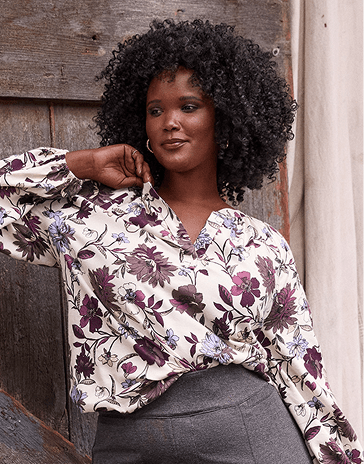 A woman with curly black hair wearing a floral blouse and grey trousers stands against a rustic wooden wall and a white curtain.
