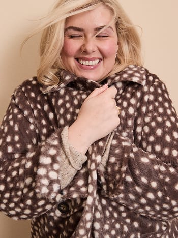 A smiling blonde woman wears a brown and white spotted faux-fur coat against a plain beige background.