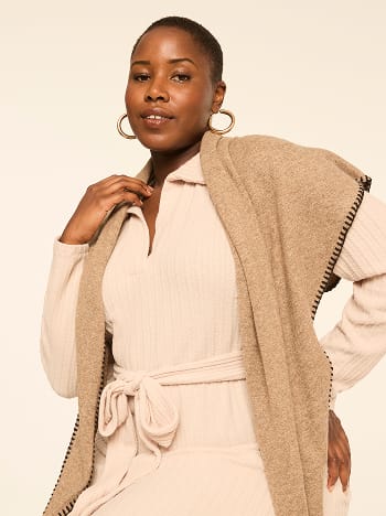 A woman wearing a beige ribbed outfit and a brown shawl stands against a plain, light-colored background.