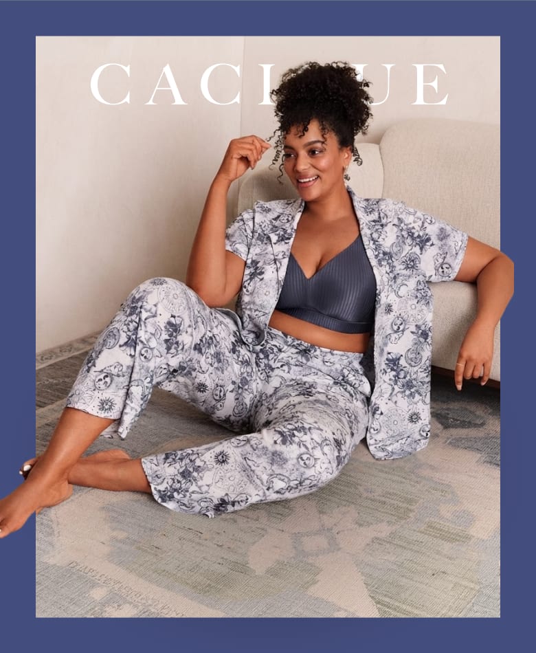 A woman wearing a grey floral pajama set and a dark blue bra sits on a patterned rug in front of a neutral-toned wall and sofa.