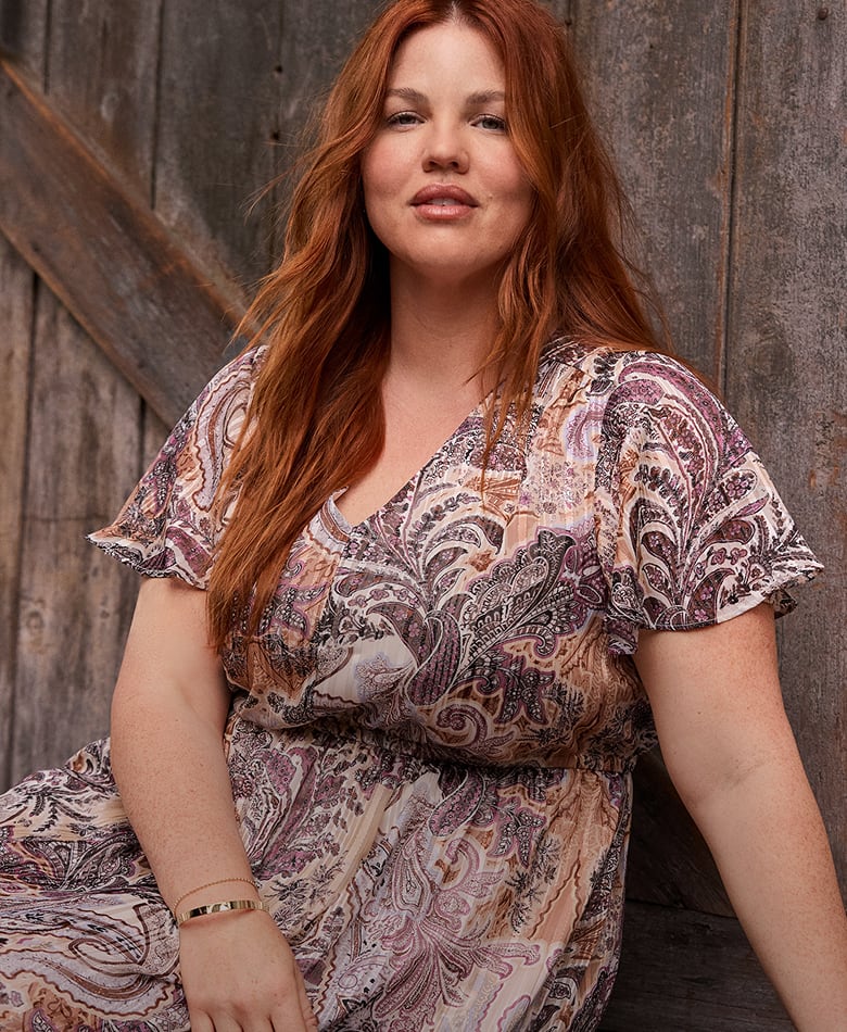 A woman with long red hair wearing a paisley print dress sits against a rustic wooden wall.