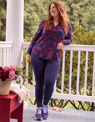 A woman in a floral top and navy leggings leans against a white porch railing with a red table and flowers in the foreground and lush green trees in the background.