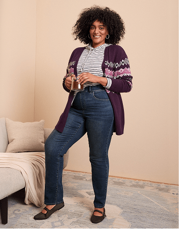 A smiling woman with curly hair wearing a purple cardigan and blue jeans stands in a room with a neutral-colored wall and a beige sofa.