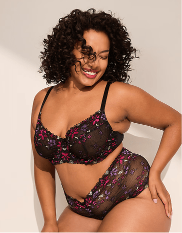 A smiling woman with curly hair wears black floral lingerie against a plain, light-colored background.