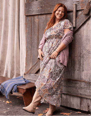 A smiling woman with red hair wearing a patterned dress and purple cardigan leans against a rustic wooden barn door next to a bench with a blue blanket.