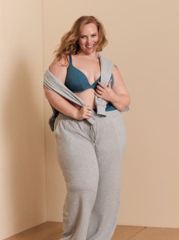 A smiling woman wearing a blue bra and grey lounge pants stands against a plain, neutral-toned wall.