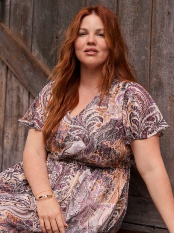 A woman with long red hair wearing a paisley print dress leans against a rustic wooden wall.