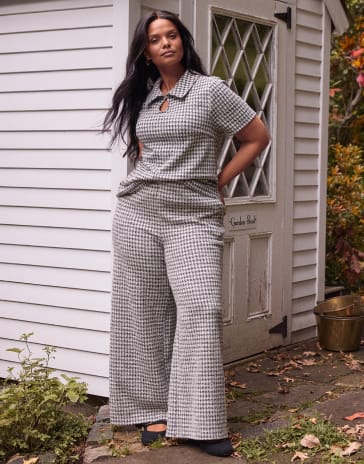 A woman wearing a grey and white patterned two-piece set stands in front of a white wooden shed and a stone path.