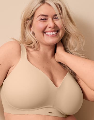 A smiling blonde woman wearing a beige bra poses against a plain, neutral-colored background.