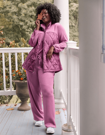 A woman in a purple quilted jacket and matching pants stands on a white porch with a lush green forest in the background.