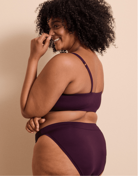 A smiling woman with curly hair wearing a dark purple two-piece swimsuit poses against a plain, light beige background.