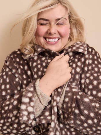 A smiling blonde woman wears a brown and white spotted faux-fur coat against a plain beige background.