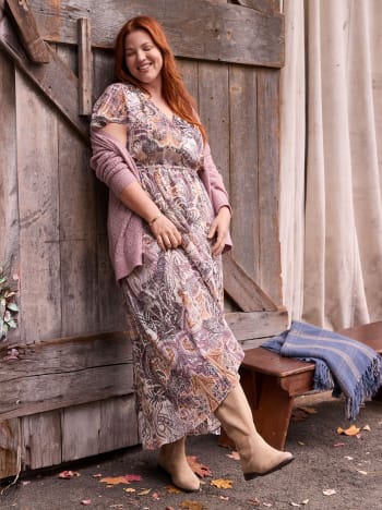 A woman with long red hair wearing a paisley print dress leans against a rustic wooden wall.