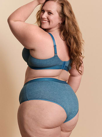 A smiling woman with long red hair wears a blue lingerie set while posing against a plain beige background.