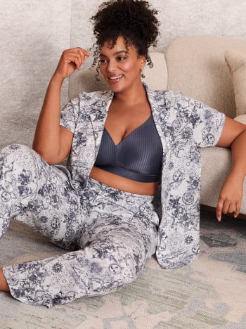 A smiling woman wearing a blue bra and grey lounge pants stands against a plain, neutral-toned wall.