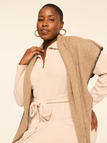A woman wearing a beige ribbed outfit and a brown shawl stands against a plain, light-colored background.