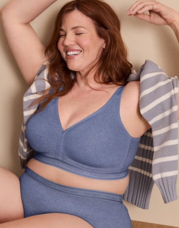 A smiling woman with red hair wears a blue bra and underwear set with a striped cardigan draped over her shoulders against a plain beige background.