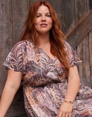 A woman with long red hair wearing a paisley print dress sits against a rustic wooden wall.