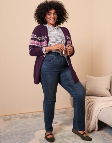 A woman wearing a purple cardigan and blue jeans stands in a room with a neutral wall and a beige sofa.