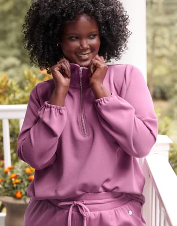 A woman in a purple quilted jacket and matching pants stands on a white porch with a lush green forest in the background.