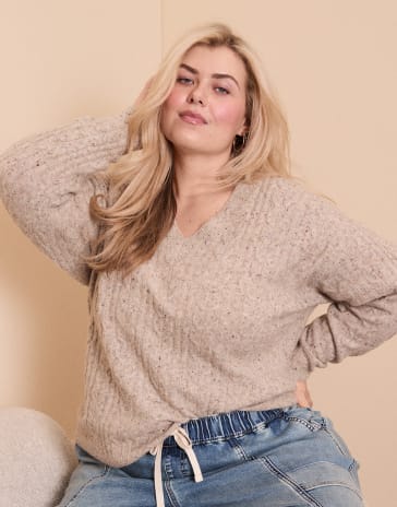 A blonde woman wearing a beige knit sweater and blue jeans poses against a plain, neutral-colored wall.