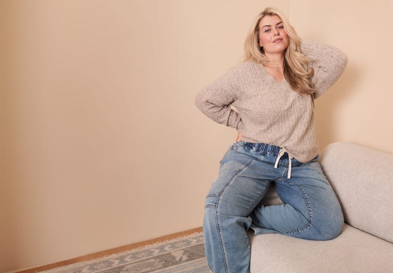 A blonde woman wearing a beige sweater and blue jeans sits on a light-colored sofa against a plain beige wall.