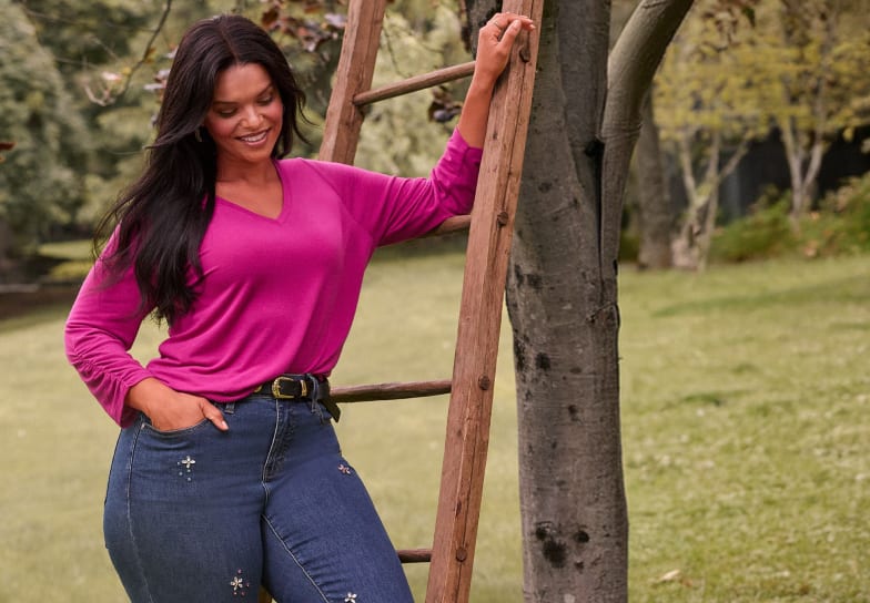 A smiling woman in a magenta v-neck top and blue jeans leans against a wooden ladder in a grassy park with trees.