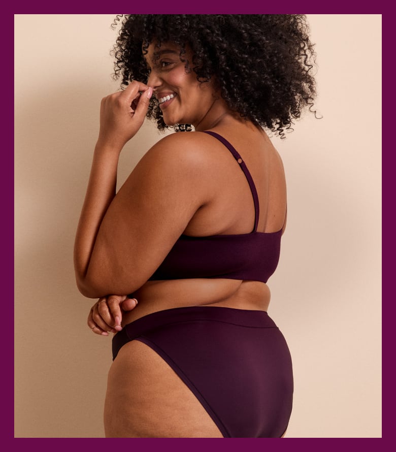 A smiling woman with curly hair wearing a dark purple lingerie set poses against a plain beige background.