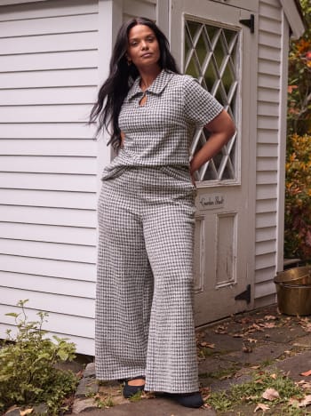 A woman wearing a grey and white checkered matching set stands in front of a white wooden house with a lattice door.