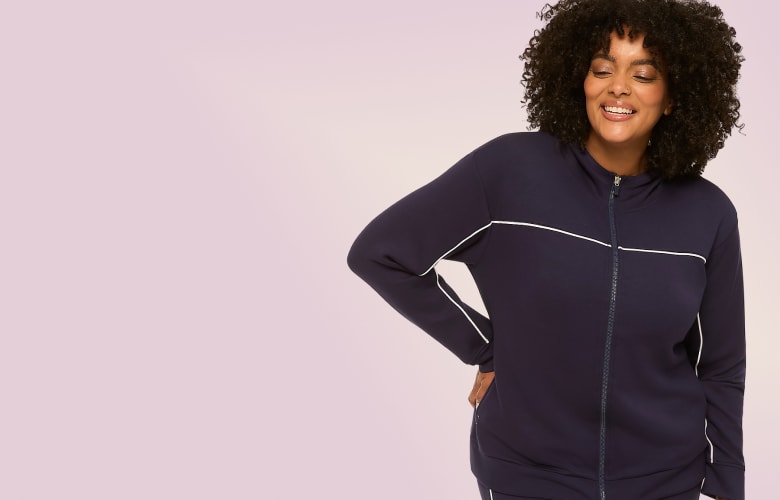 A smiling woman with curly hair wearing a navy blue zip-up jacket stands against a soft, gradient purple and white background.