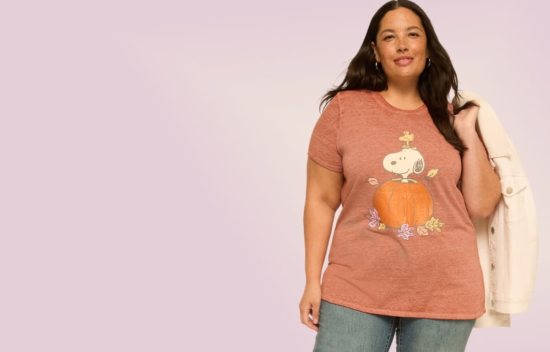 A woman wearing a rust-colored Snoopy t-shirt and jeans stands against a plain, light purple background.