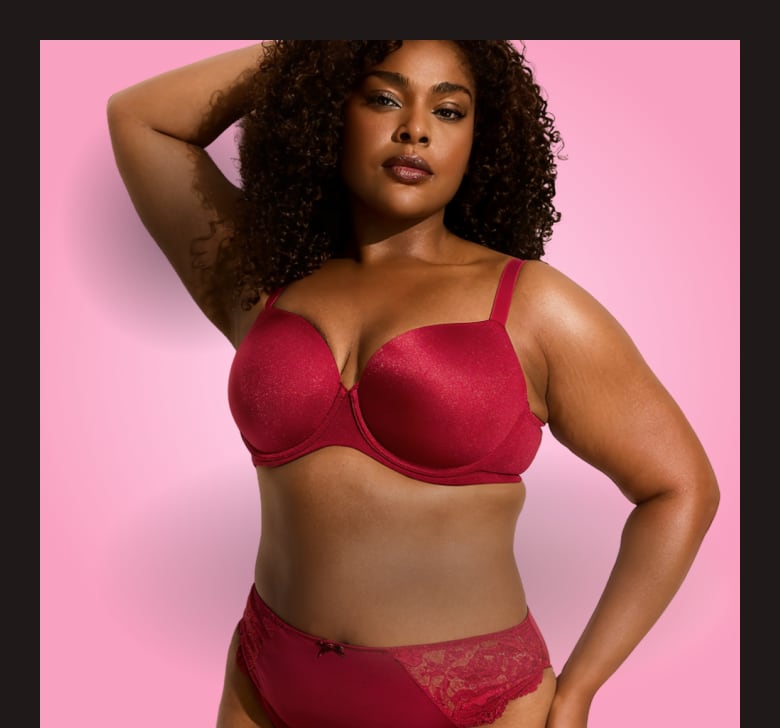 A woman with curly hair wearing a red lingerie set poses against a solid pink background.