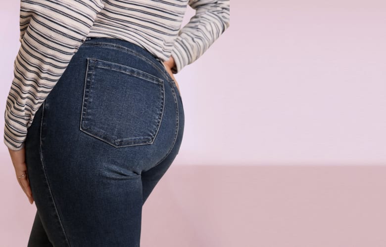 A person wearing a striped shirt and dark blue jeans stands against a plain, light pink background.
