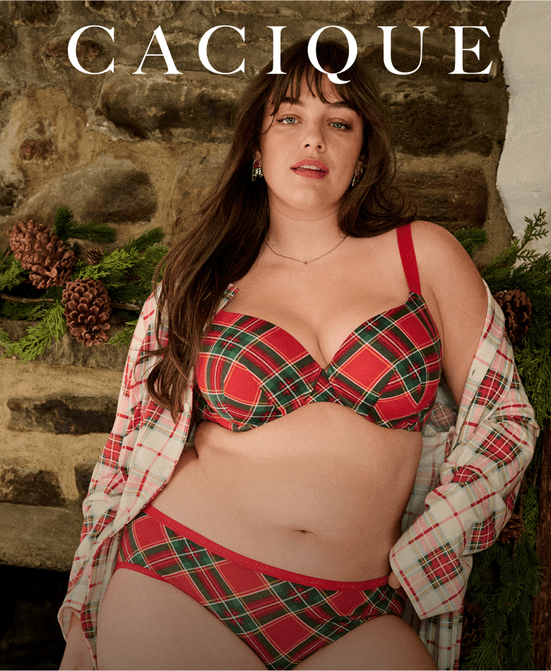 A woman wearing red and green plaid lingerie and an open matching shirt poses in front of a rustic stone fireplace decorated with pine branches and pinecones.