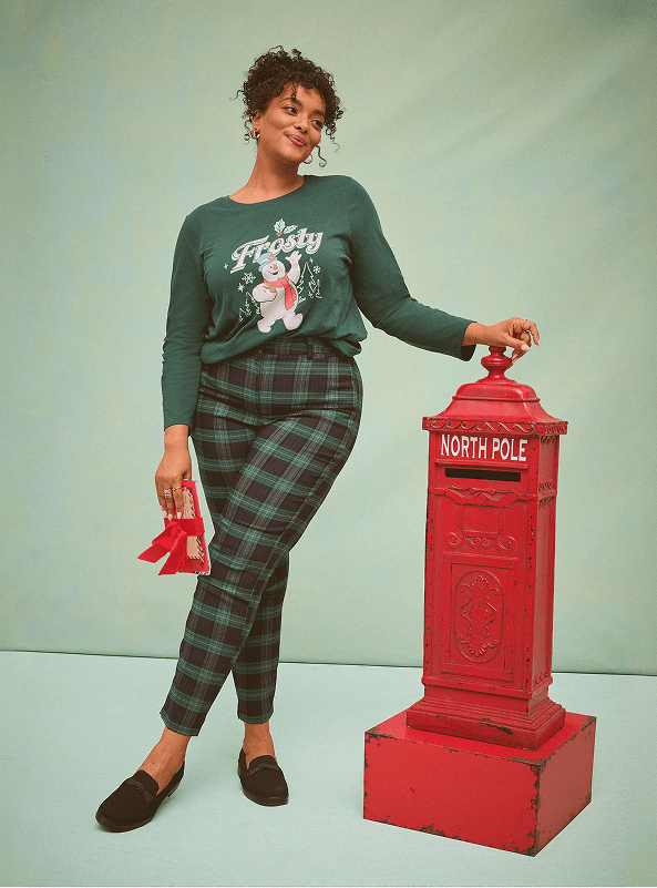A woman wearing a green 'Frosty' sweater and plaid pants leans against a red North Pole mailbox against a plain light-blue background.