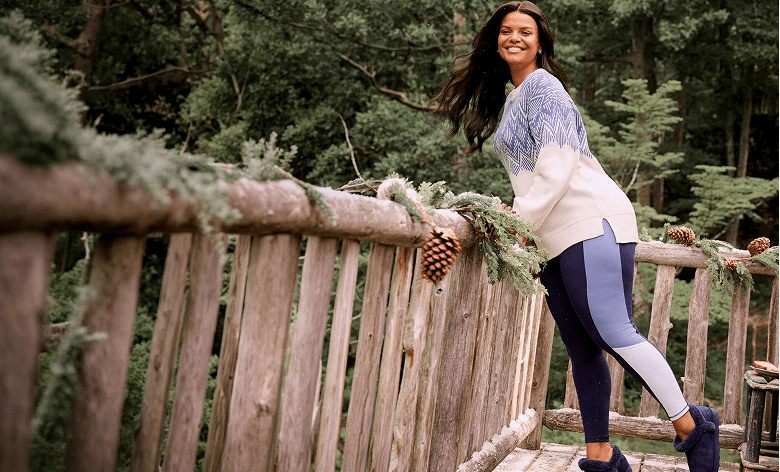 A smiling woman in a patterned sweater and leggings leans against a rustic wooden railing decorated with pine branches, set against a dense forest background.