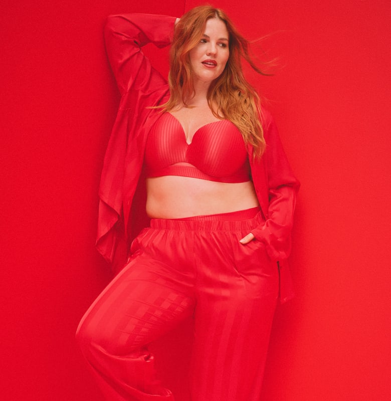 A woman wearing a red bra, satin pants, and a matching robe poses against a solid red background.