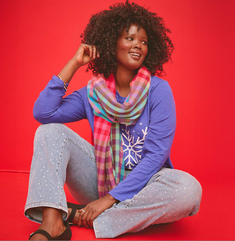 A woman with curly hair wearing a purple shirt, colorful scarf, and patterned jeans sits against a solid red background.