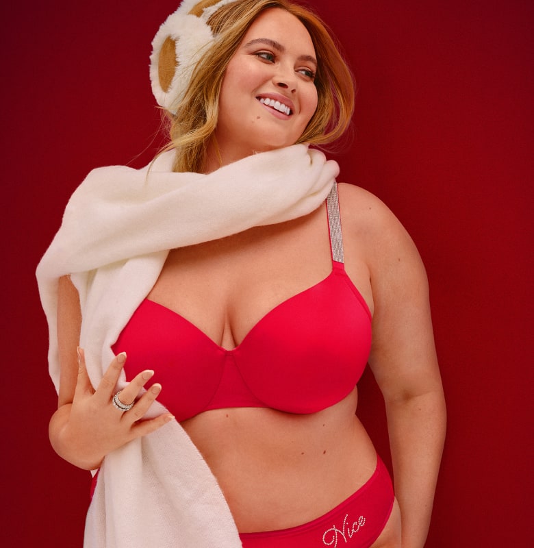 A smiling woman wearing red lingerie, a white fluffy wrap, and a fuzzy hat poses against a solid red background.