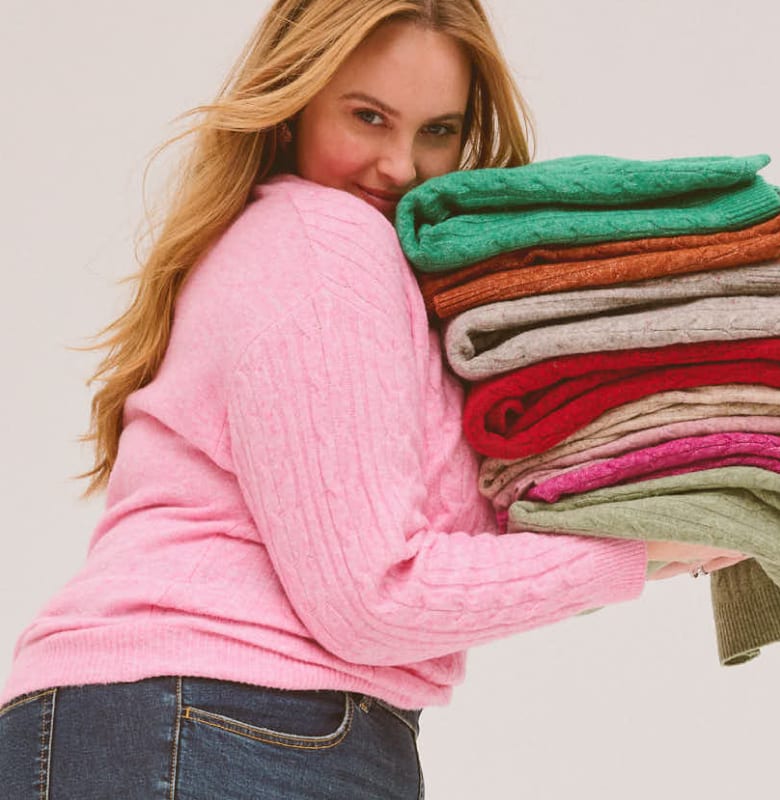 A woman in a pink sweater holds a colorful stack of folded sweaters against a plain white background.