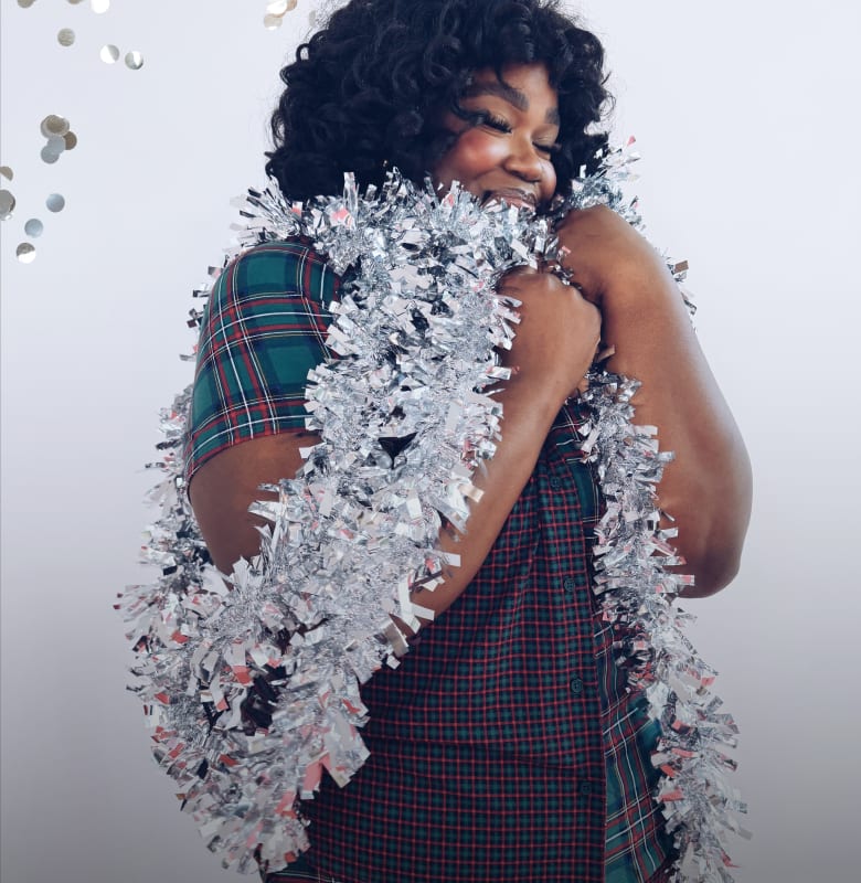 Three women pose in separate panels against a plain white background, featuring one wrapped in silver tinsel, one holding a disco ball, and one balancing a red gift on her head.