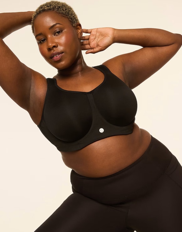A woman wearing a black sports bra and leggings poses with her arms raised against a plain, light-colored background.