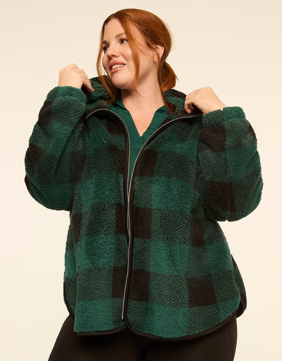 A woman with red hair wearing a green and black checkered fleece jacket stands against a plain, light-colored background.