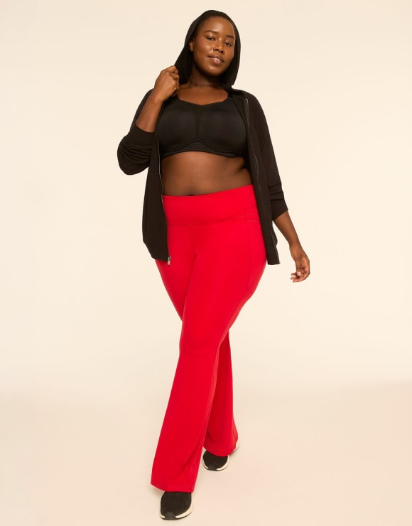 A woman wearing a black sports bra, black zip-up hoodie, and red flared pants stands against a plain, light-colored background.