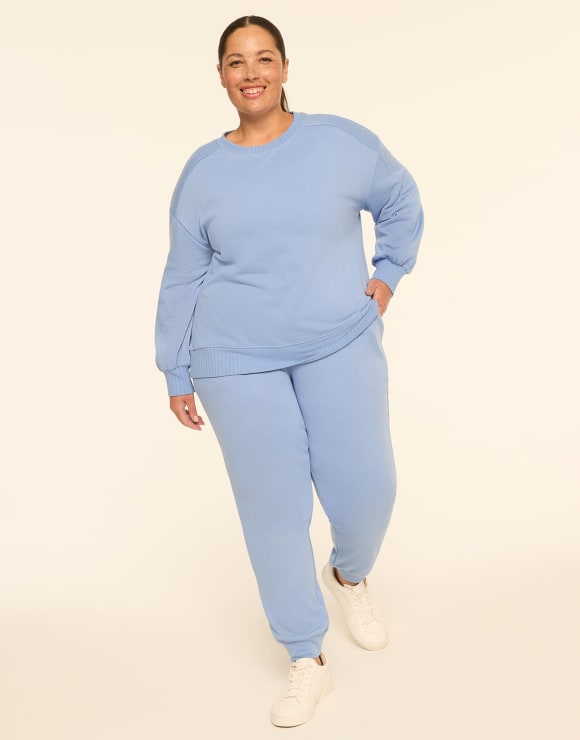 A smiling woman wearing a light blue sweatshirt and matching sweatpants stands against a plain, off-white background.